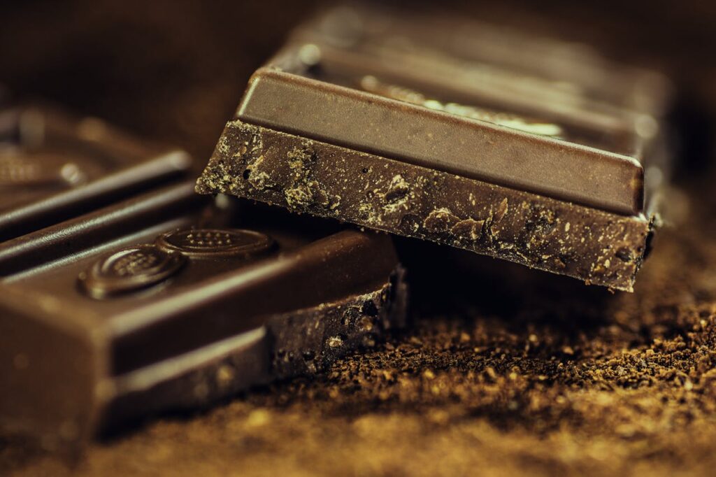 Broken dark chocolate bars stacked on cocoa powder, showing rich texture and crumbly interior in close-up detail.